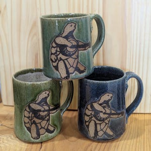 Puede incluir: Tres tazas de cerámica apiladas, en tonos verdes y azules. Cada taza presenta un diseño de tortuga marrón. Las tazas tienen un acabado brillante y un asa curvada. Adecuadas para bebidas frías o calientes.