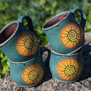 May include: Four turquoise ceramic mugs stacked together. Each mug features a hand-painted sun design in orange and yellow. The interior of the mugs is a deep red color, and the mugs have a speckled texture.