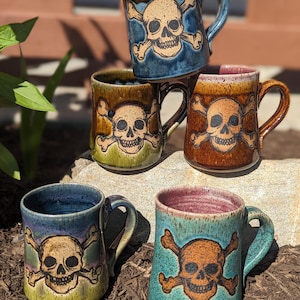 Könnte beinhalten: Eine Sammlung handgefertigter Keramikbecher mit Totenkopf- und gekreuzten Knochen-Design. Die Becher sind in verschiedenen Farben erhältlich, darunter Blau, Braun und Grün, mit glänzendem Finish. Jeder Becher hat einen Henkel und ein einzigartiges Glasurmuster.