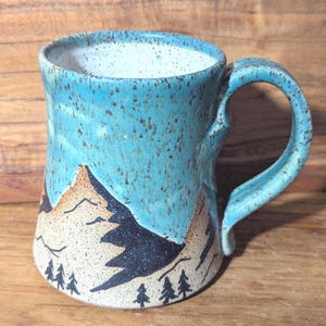 May include: A turquoise ceramic mug with a mountain scene and a speckled glaze. The mug has a brown handle and a white interior.
