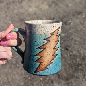 May include: A ceramic mug with a teal and tan gradient, featuring a brown lightning bolt design. The mug has a speckled finish and a curved handle. The mug is held by a person with pink nail polish.
