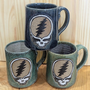 Puede incluir: Tres tazas de cerámica con el diseño Steal Your Face. Las tazas son en tonos de azul y verde, con un gráfico de calavera y rayo. Cada taza tiene un asa y un acabado texturizado.