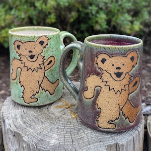 May include: Two ceramic mugs with handles, each featuring a dancing bear design. One mug is green, the other is burgundy. The bears are tan with black outlines. The mugs are sitting on a weathered wooden surface.