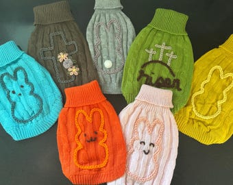 May include: Seven knitted dog sweaters in various colours with different embroidered designs. The designs include a cross, a bunny, and the word "risen".