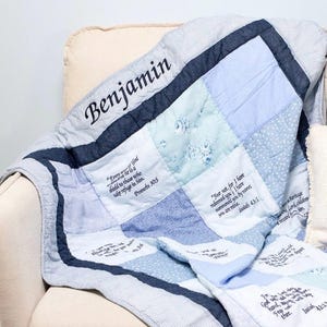 May include: A patchwork quilt in shades of blue and white, featuring the name "Benjamin" embroidered in black. The quilt has various fabric patterns and embroidered Bible verses. A cozy, handmade blanket.