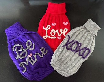 May include: Three knitted dog sweaters with different Valentine's Day themed messages. The first sweater is purple with the message "Be Mine" in gray yarn. The second sweater is red with the message "Love" in white yarn. The third sweater is gray with the message "XOXO" in purple yarn.