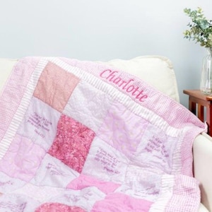 May include: A pink and white patchwork quilt with the name "Charlotte" embroidered in pink thread. The quilt features various shades of pink squares and printed text. The quilt is draped over a white couch.