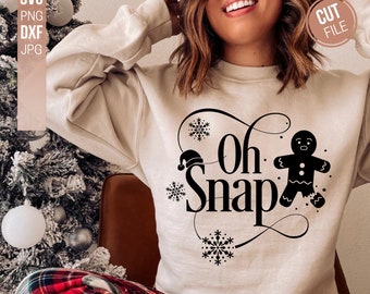 May include: A beige crewneck sweatshirt featuring a black graphic print of a gingerbread person with a missing leg, a Santa hat, and snowflakes. The text reads Oh Snap in a decorative font. The design includes SVG, PNG, DXF, and JPG file labels.