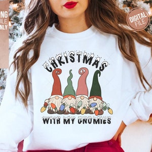 May include: A white sweatshirt featuring a graphic of four gnomes wearing red, green, and pink hats, holding colorful Christmas lights. The text above reads CHRISTMAS and the text below reads WITH MY GNOMIES in a bold, black, snowy-style font.