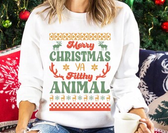 May include: A white long-sleeve sweatshirt featuring the text Merry Christmas Ya Filthy Animal in red and green. The design includes graphic elements of reindeer, snowflakes, and a checkered pattern in orange, red, and green colors.