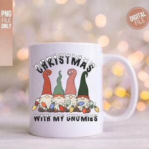 May include: A white ceramic mug featuring a graphic of four gnomes wearing red, green, and pink hats, holding colorful Christmas lights. The text above reads CHRISTMAS and the text below reads WITH MY GNOMIES in black, snow-capped lettering.
