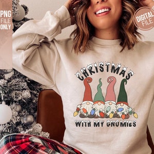May include: A beige crewneck sweatshirt featuring a graphic of four gnomes wearing tall hats in red, green, and pink. The gnomes hold colorful Christmas light bulbs. The text reads CHRISTMAS WITH MY GNOMIES in black with white snow accents.