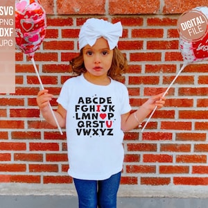 May include: A white t-shirt with black letters spelling out the alphabet. The letter "I" is replaced with a red heart and the letter "U" is replaced with a red heart. The shirt is being worn by a child holding a red heart balloon.