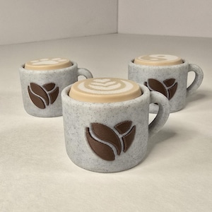 Może przedstawiać: Trzy małe, nakrapiane szare kubki do kawy z brązowymi wzorami ziaren kawy. Każdy kubek ma pokrywkę w kolorze beżowym, w stylu latte art. Kubki są ułożone na białej powierzchni, co sugeruje produkt o tematyce kawowej.