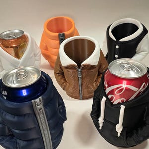 May include: A collection of beverage can holders shaped like jackets. The holders are in various colors, including orange, navy blue, brown, black, and white. Each holder has a zipper or button details, and the cans are visible inside.