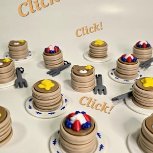 May include: An assortment of miniature pancake-shaped clickers, some topped with butter, others with berries and whipped cream. Each sits on a small plate with a blue floral design. The words "Pancake Clickers!" and "Click!" are displayed.