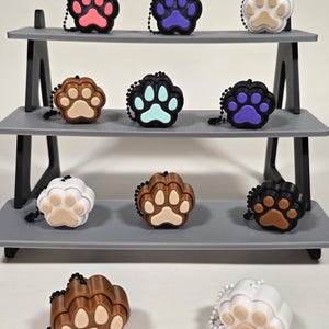 Pode incluir: Uma exibição de chaveiros em forma de pata em várias cores, incluindo rosa, roxo, castanho e branco. Cada chaveiro apresenta um design de pegada contrastante e uma pequena corrente. Os chaveiros estão dispostos numa prateleira cinzenta de três níveis.