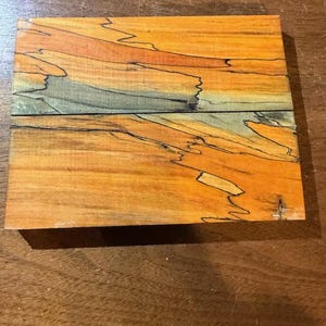May include: Two rectangular pieces of wood with a vibrant, natural grain pattern. The wood features a warm, orange-yellow base color with streaks of orange, blue-green, and black, creating a unique, artistic appearance. The wood is likely for crafting or woodworking projects.