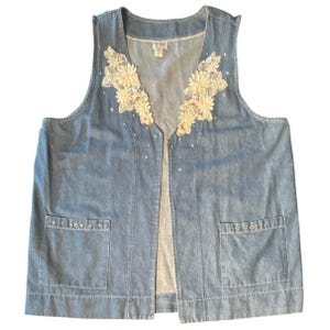 May include: A sleeveless blue denim vest featuring a V-neckline adorned with cream-colored floral lace appliqués and small clear rhinestones. The garment includes two front patch pockets, each embellished with a row of small clear rhinestones.