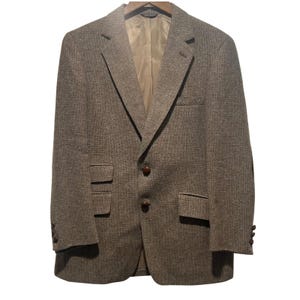 May include: A brown tweed blazer with a notched lapel, two front pockets, and a single chest pocket. The blazer has two brown buttons and three buttons on each cuff. The interior lining is a light tan colour. The blazer is hanging on a wooden hanger.