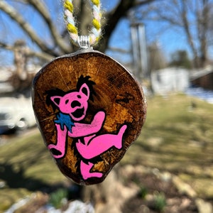 May include: A pink hand-painted bear on a wooden pendant, hanging from a yellow and white braided cord. The pendant is shaped like a tree slice.