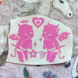 May include: A pink sticker featuring two cherubic figures, one with angel wings and a halo, the other with devil wings and horns. A heart, star, and sparkles are also included in the design.