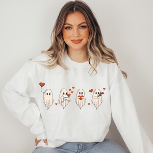 May include: White sweatshirt with a graphic of four ghosts holding hearts, balloons, and flowers. The ghosts are wearing red hearts and the text "Love You" is on one of the ghosts.