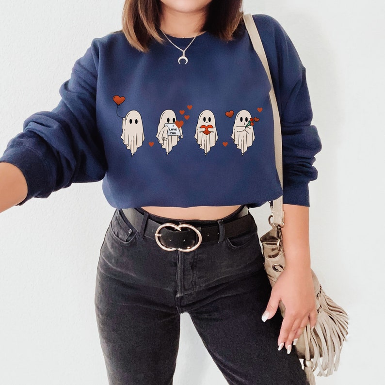 May include: A navy blue cropped sweatshirt with a graphic of four ghosts holding hearts and flowers. The text "Love You" is on one of the ghosts. The sweatshirt is worn by a person with black jeans and a gold belt.