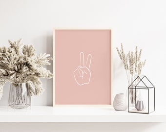 Op de afbeelding: Een ingelijst print met een roze achtergrond en een witte omtrek van een hand die het vredesteken maakt.