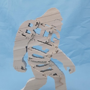 May include: A wooden jigsaw puzzle of a Bigfoot, or Sasquatch, walking. The puzzle is made of natural wood and has a light brown color.