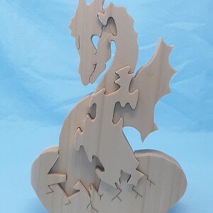 Op de afbeelding: Een houten puzzel van een draak die uit een gebarsten ei komt. De draak is gemaakt van meerdere in elkaar grijpende stukken en staat met gespreide vleugels. Het ei is één stuk met een gebarsten oppervlak.