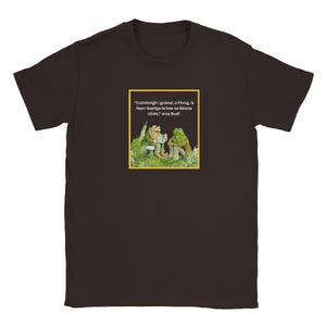 May include: A dark brown short-sleeved t-shirt featuring a square graphic of two frogs sitting in grass. One frog reads a book while the other listens. The text reads: "Cuimhnigh i gcónaí, a Fhrog, is fearr Gaeilge briste ná Béarla cliste," arsa Buaf.