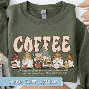 May include: Green sweatshirt with a graphic of gnomes holding coffee mugs and the text "COFFEE" in a brown and beige font. The text below the graphic reads "a Magical substance that turns the words "Leave me alone" into "Good Morning". The bottom of the sweatshirt has the text "more color options".