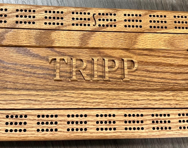 May include: A wooden cribbage board with a personalized inscription that reads "TRIPP". The board has a brown wood grain finish and features rows of holes for scoring.