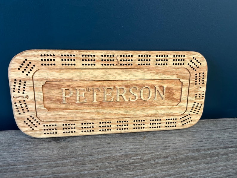 May include: A wooden cribbage board with a personalized name "PETERSON" engraved on the surface. The board has a traditional design with holes for pegs and a scoring area.