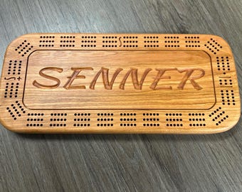 May include: A wooden cribbage board with a personalized inscription that reads "SENNER". The board has a brown stain and features a traditional cribbage board design with holes for pegs.