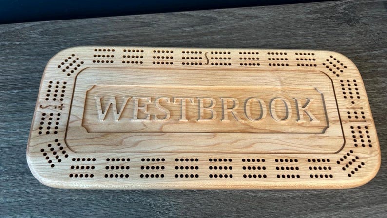 May include: A wooden cribbage board with a personalized inscription that reads "WESTBROOK". The board has a light brown color and features a traditional design with holes for pegs.