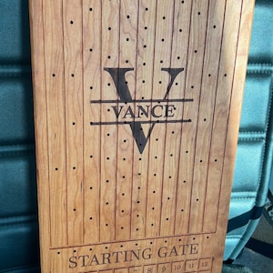 May include: A wooden board with a horse racing theme. The board has a finish line, a starting gate, and a grid of holes. The board is personalized with the name "VANCE".
