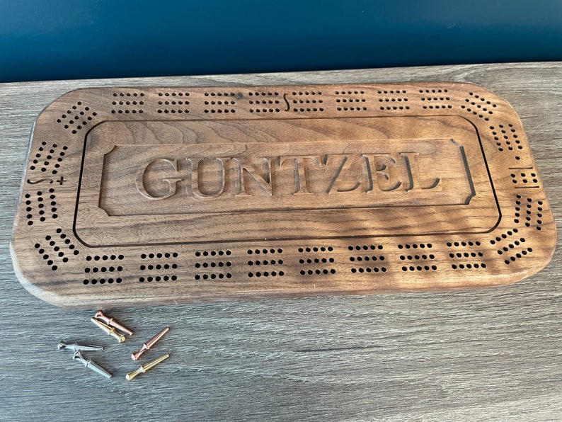 May include: A wooden cribbage board with the name "GUNTZEL" engraved on the top. The board has a dark brown stain and is made of walnut wood. The board is surrounded by a border of small holes.
