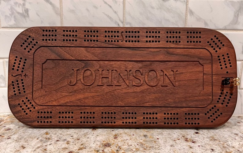 May include: A personalized wooden cribbage board with the name "JOHNSON" engraved on the surface. The board has a dark brown walnut finish and features a traditional cribbage board design with holes for pegs.