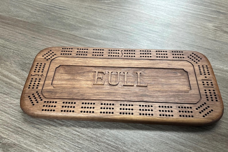 May include: A wooden cribbage board with a personalized inscription that reads "FULL". The board has a dark brown stain and features a traditional cribbage board design with holes for pegs.