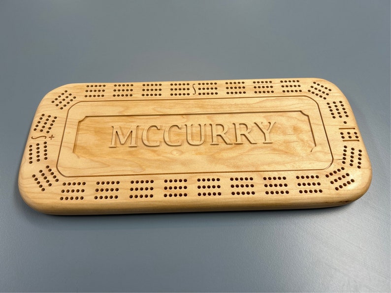 May include: A wooden cribbage board with the name "MCCURRY" engraved on the surface. The board has a traditional design with holes for pegs and a scoring area.