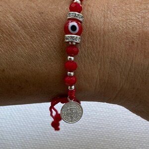 May include: Red beaded bracelet with a silver charm and a red evil eye bead. The charm has the words "Sanctus Paulus" and a cross.