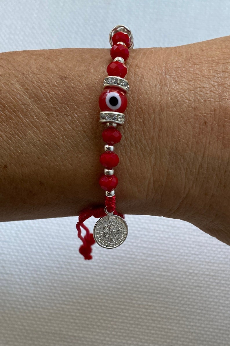 May include: Red beaded bracelet with a silver charm and a red evil eye bead. The charm has the words "Sanctus Paulus" and a cross.