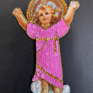 May include: A pink sequined religious figure of a young child with a halo, standing on a white sequined cloud. The figure is made of fabric and sequins and is a traditional religious art piece.