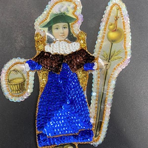 May include: A colorful, sequined appliqué of a young girl holding a basket and a staff with a golden orb. The girl is wearing a blue dress with a brown cape and a white hat. The appliqué is made of sequins, beads, and fabric.