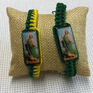 May include: Two green and yellow braided bracelets with a rectangular image of a saint holding a staff and a book. The image is on a white background with a gold frame.