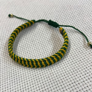 May include: A green and yellow braided bracelet with adjustable length. The bracelet is made with two colors of cord and has a simple knot closure.