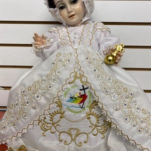 May include: A white doll wearing a white satin dress with gold embroidery and a matching bonnet. The dress features a detailed design with pearls and the text "Jubileo 2025". The doll holds a gold cross and orb.