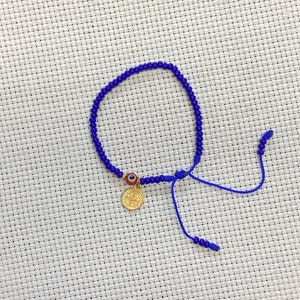 May include: A blue beaded bracelet with a gold charm featuring an evil eye design. The bracelet is tied with a blue cord.